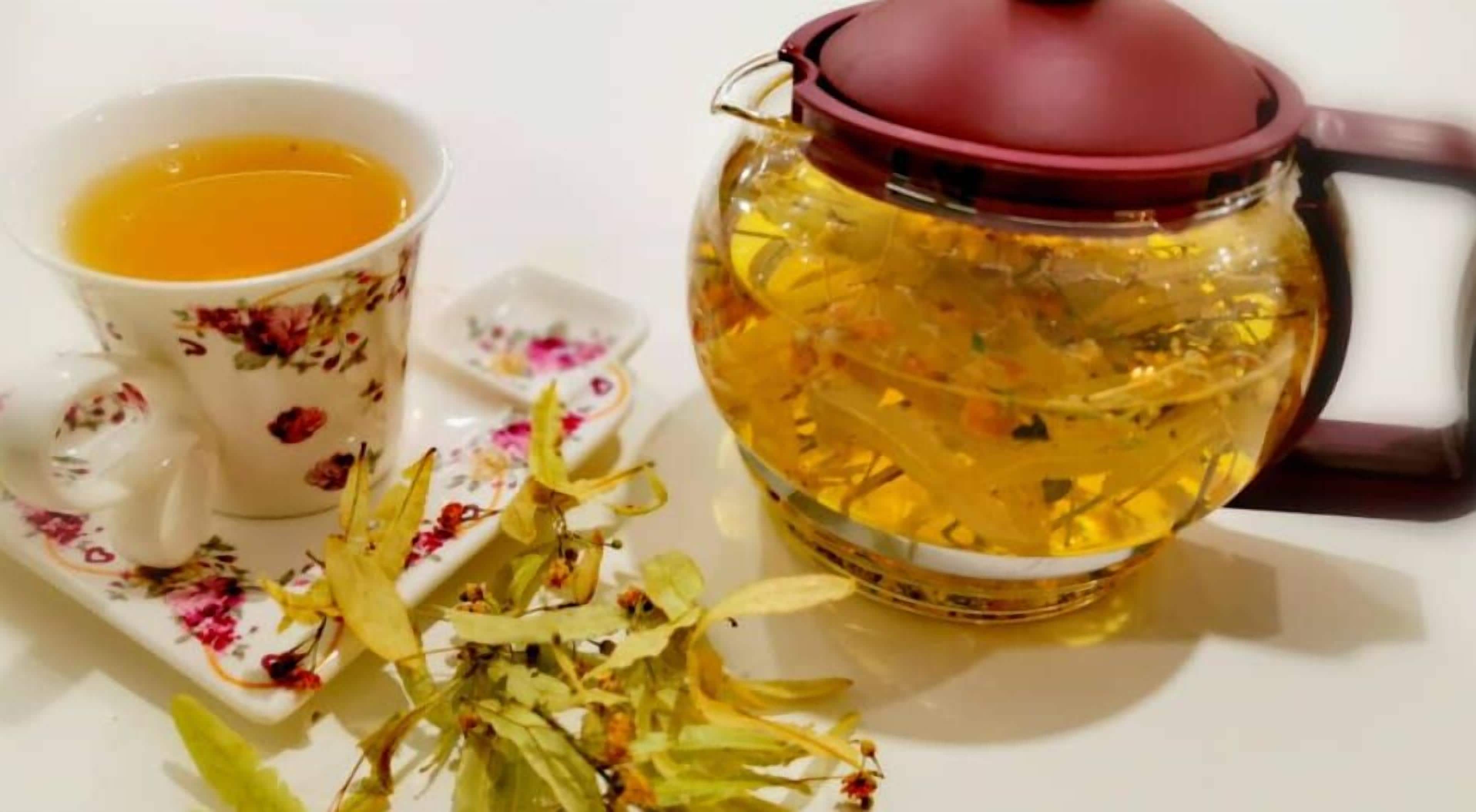Una tetera y una taza con diseños florales, sobre una superficie blanca con hojas y tallos de té.