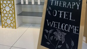 Exhibició de caixes de la colònia LAGUS sobre prestatges, un cartell que diu 'ROSE THERAPY HOTEL WELCOME' i una paret decorativa amb un dibuix geomètric.