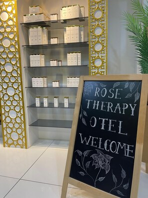 A display of LAGUS cologne boxes on shelves, a sign reading 'ROSE THERAPY HOTEL WELCOME,' and a decorative wall with a geometric pattern. - Rosa Therapy Hotel (Isparta)