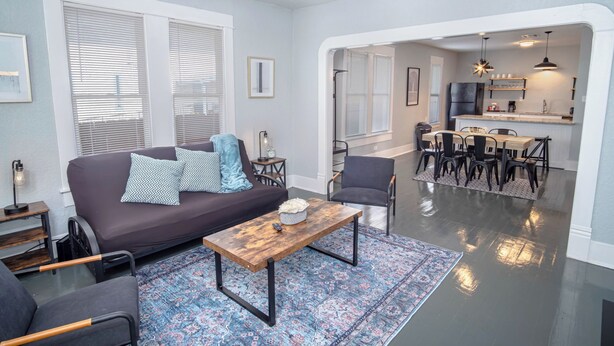 Una sala de estar moderna con un sofá, una mesa de centro y sillas, que da a un comedor con una cocina al fondo.