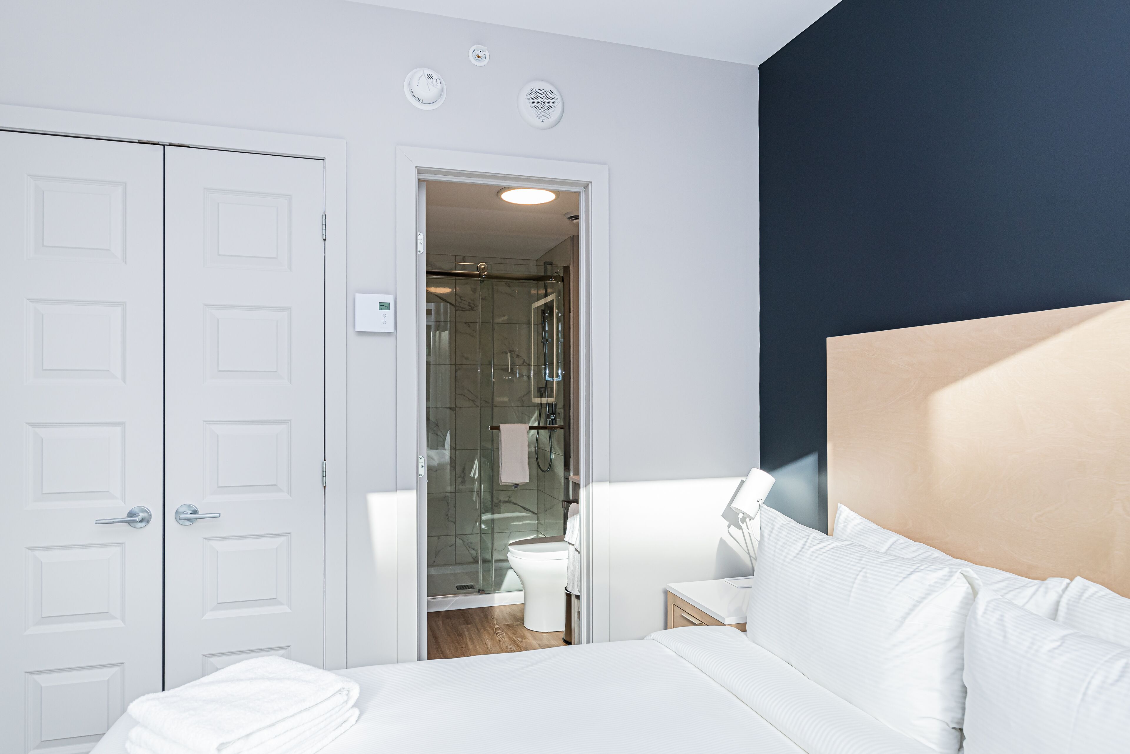 A hotel room with a bed, a bedside table, a nightstand, a white wardrobe, and a bathroom visible through an open door.