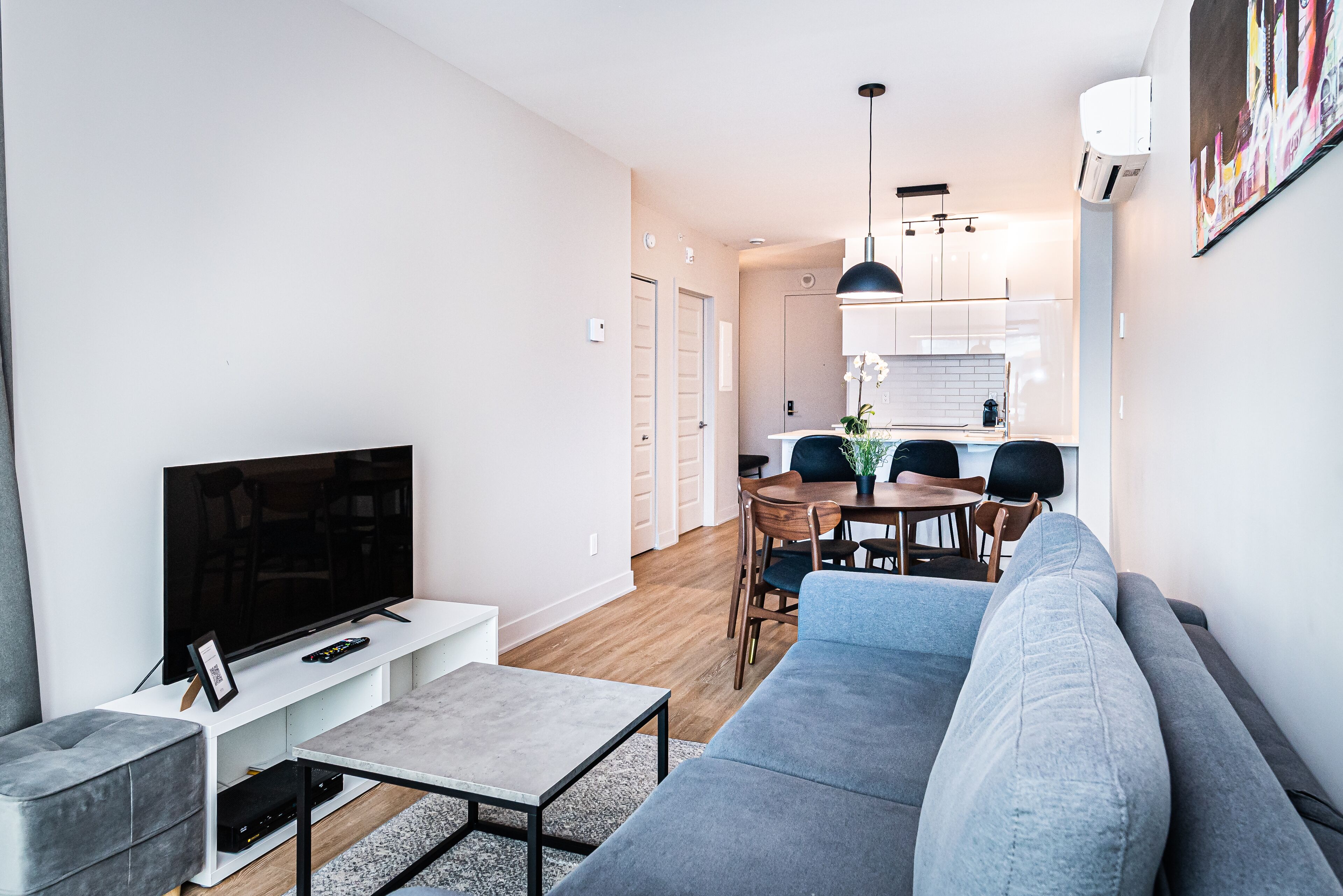Una sala de estar moderna con un sofá gris, un televisor de pantalla plana y una zona de comedor con una mesa y sillas de madera.