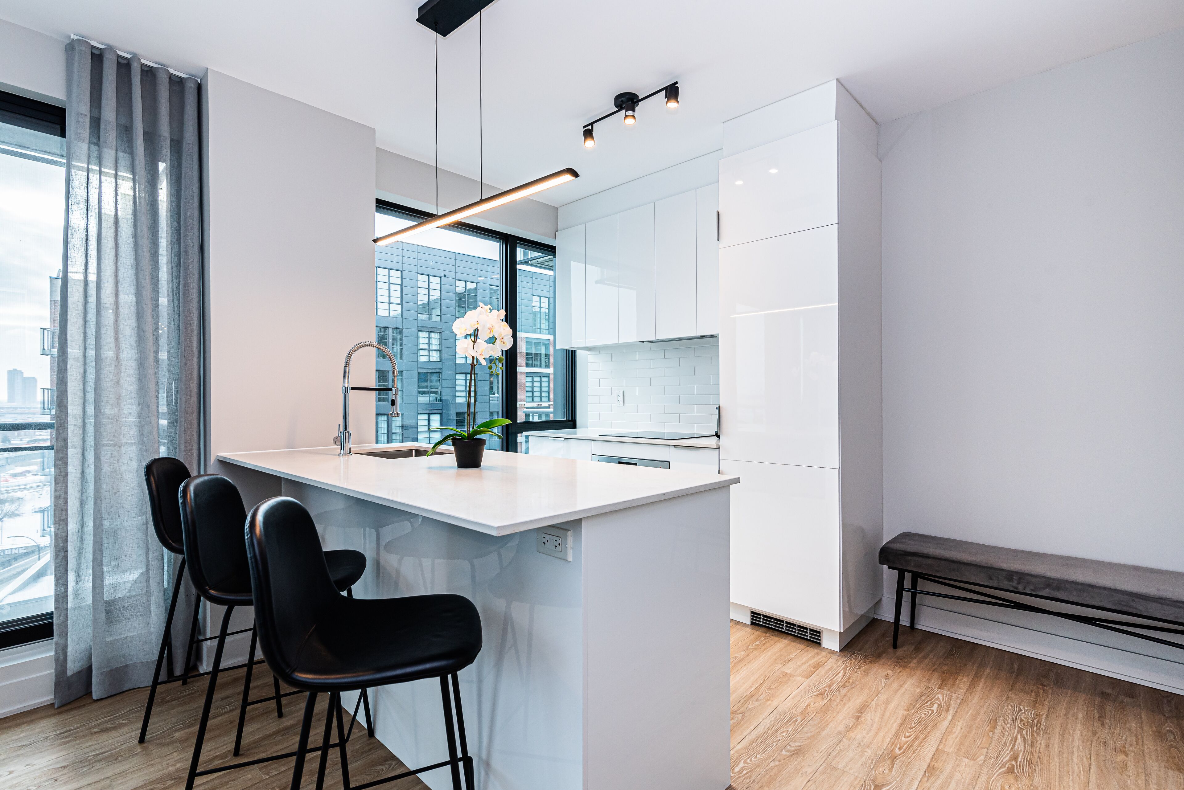 A modern kitchen with white cabinetry, a central island with bar stools, a bench, and a view of the city through large windows.