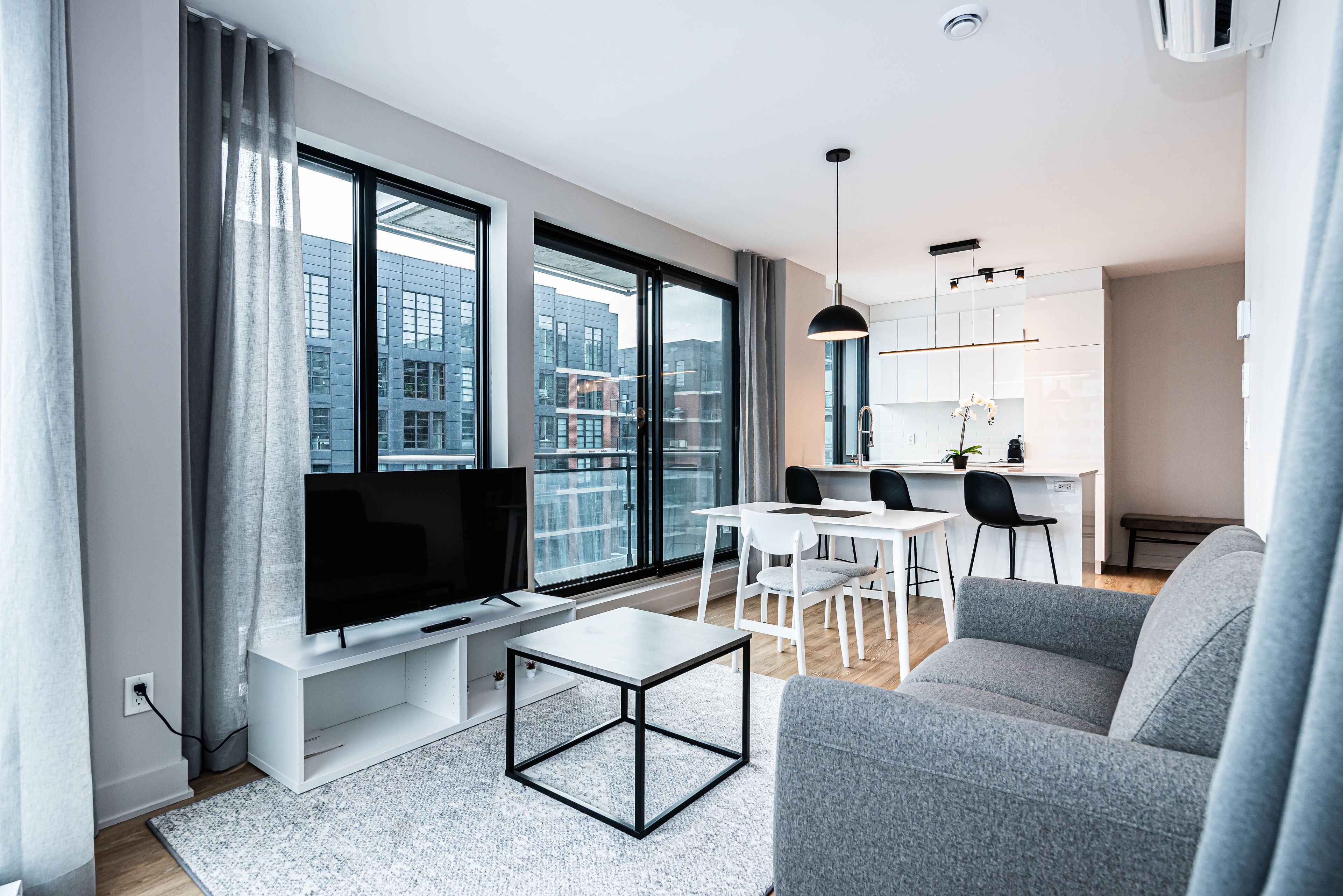 Een moderne woonkamer met een flatscreen televisie, een grijze bank en een eethoek met witte meubels.