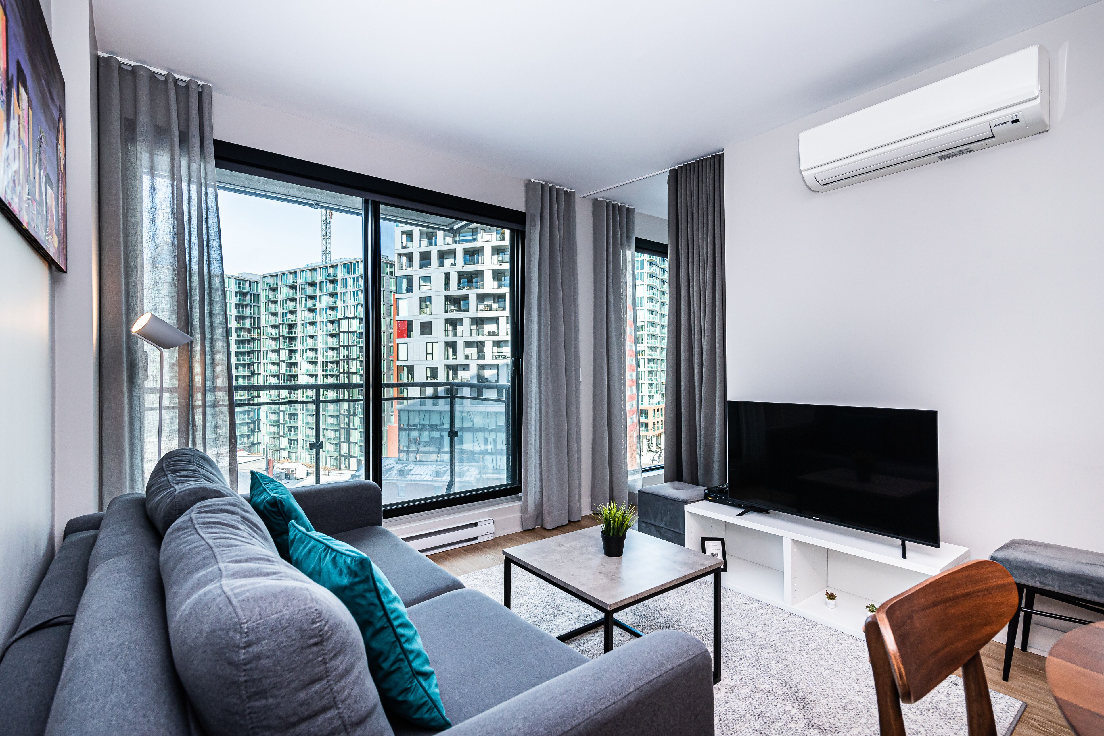 A modern living room with a large window, grey sofa, flat-screen TV, and a small table.