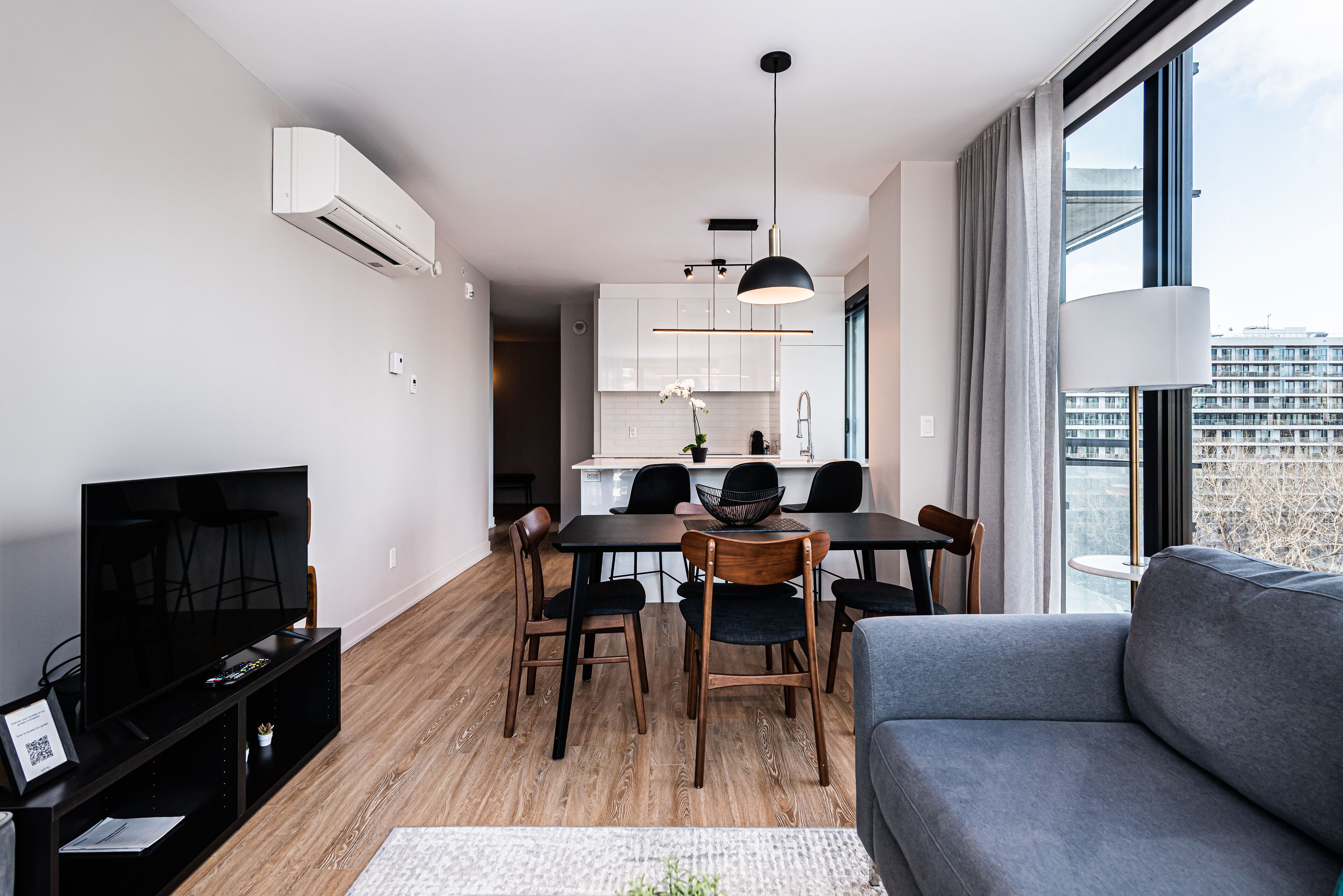 A modern living room with a flat-screen TV, a grey sofa, and a dining area with wooden chairs and a black table.