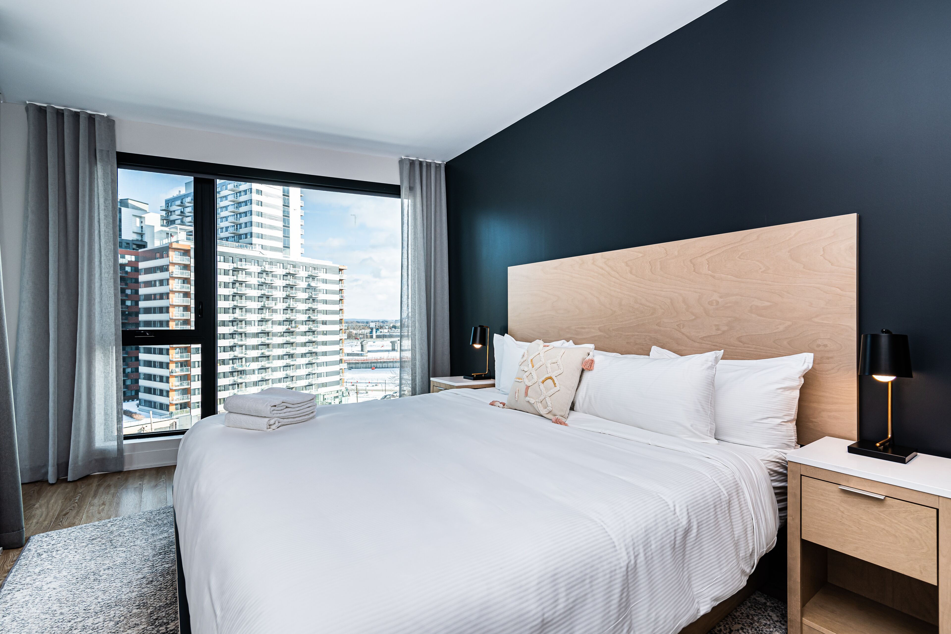 A modern hotel room with a large bed, a wooden headboard, a nightstand with a lamp, and a view of tall buildings through a large window.