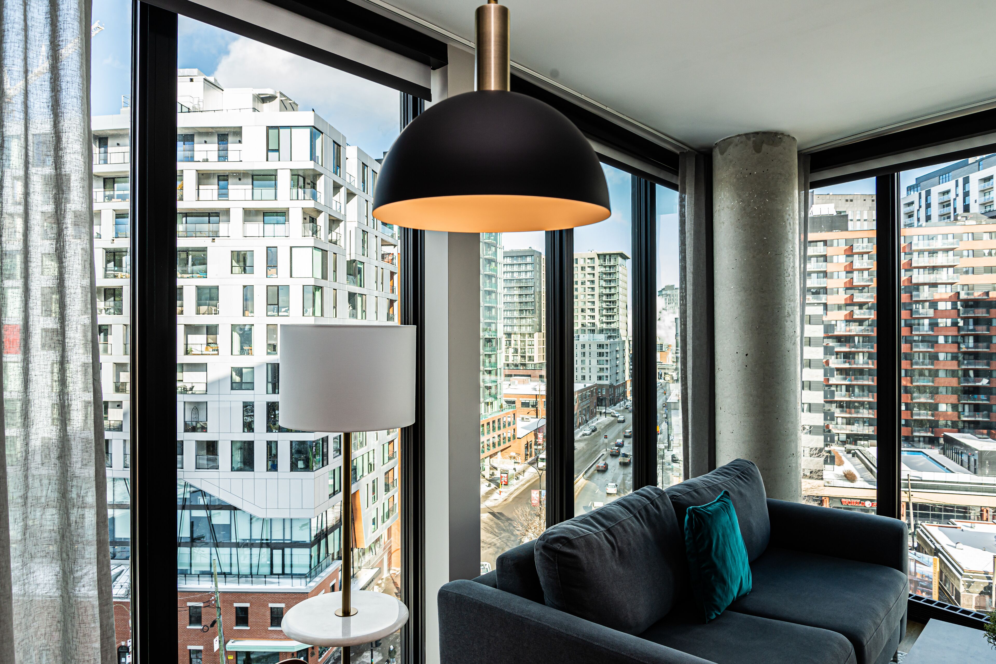 A modern living room with a large window, a black pendant lamp, and a grey sofa with a teal pillow.