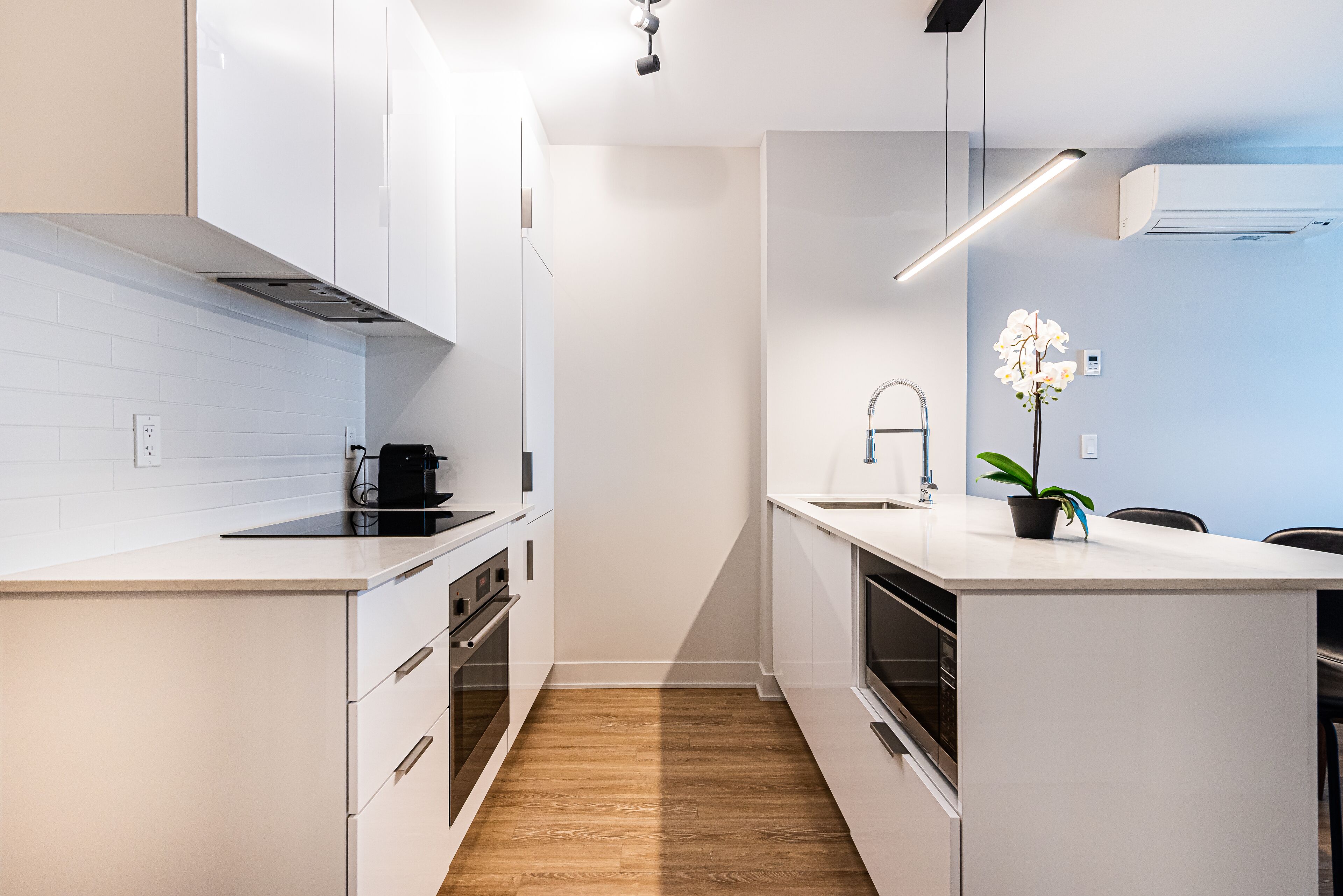 Une cuisine moderne avec des meubles blancs, un plancher en bois et un petit îlot central doté d’un évier et d’une orchidée en pot.