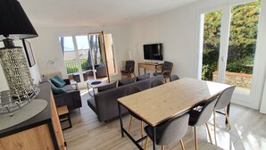A modern living room with a sofa set, a dining table with chairs, a TV, and a large window with a view of trees.