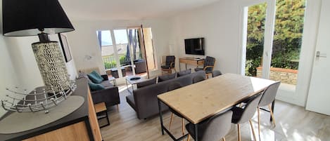 A modern living room with a sofa set, a dining table with chairs, a TV, and a large window with a view of trees.