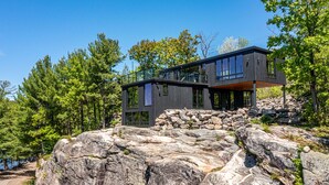 A modern two-story house built on a rocky hillside with large windows and a stone foundation. - Modern 4 bd, 3 bath Cottage on Boshkung Lake, Haliburton (Algonquin Highlands)