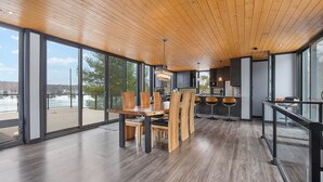 A spacious dining area with a large wooden table and chairs, a chandelier, and large windows offering a view of the outdoors. - Modern 4 bd, 3 bath Cottage on Boshkung Lake, Haliburton (Algonquin Highlands)