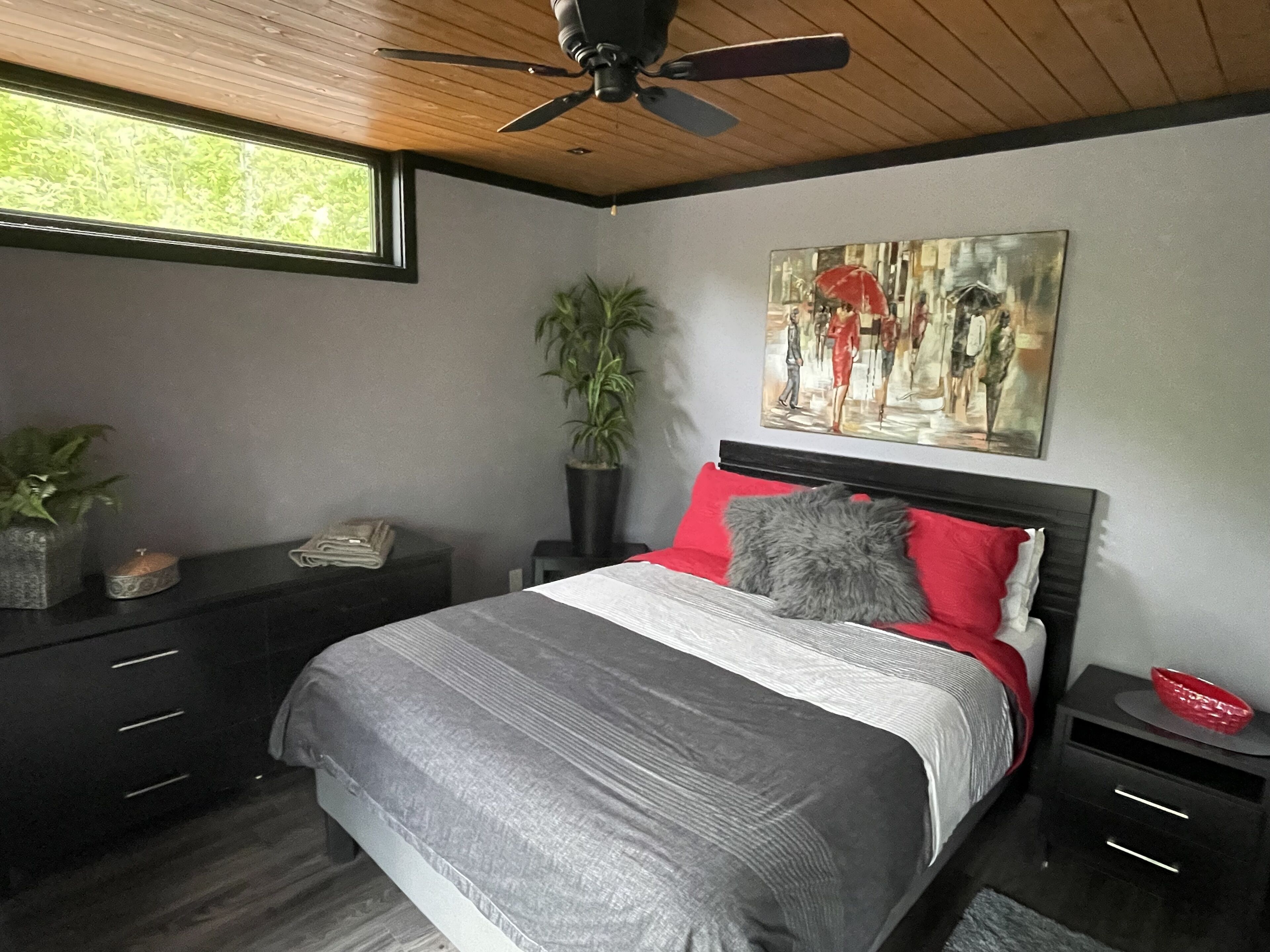 Un dormitorio con cama, mesita de noche, planta en maceta, un cuadro y ventilador de techo.