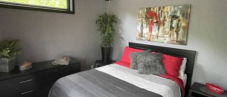 Un dormitorio con cama, mesita de noche, planta en maceta, un cuadro y ventilador de techo.