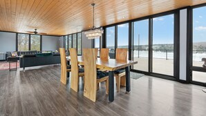 A spacious dining area with a wooden table and chairs, large windows offering a view of a body of water, and a ceiling fan. - Modern 4 bd, 3 bath Cottage on Boshkung Lake, Haliburton (Algonquin Highlands)