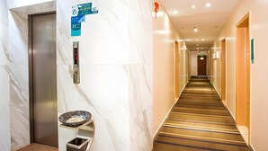 A hotel hallway with a marble wall, an elevator, a trash bin, and a sign indicating amenities. - City Comfort Inn (Hezhou)
