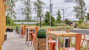 An outdoor seating area with wooden tables and orange chairs, surrounded by trees and a view of a body of water. - Premier Inn Milton Keynes - Willen Lake (Milton Keynes)
