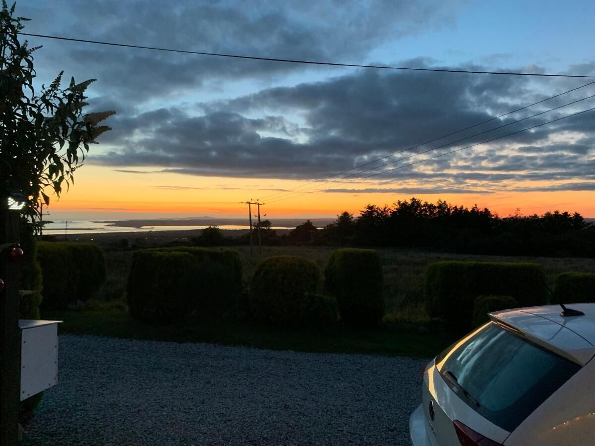 A residential area with a car parked in the foreground, a gravel driveway, and a view of the sunset over a body of water with trees and bushes in the background.
