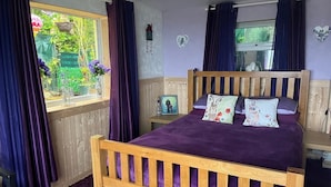 Un dormitorio con una cama de madera, ropa de cama morada, una ventana con cortinas y una mesita de noche pequeña con un marco de fotos.