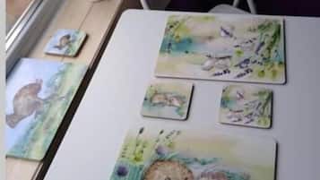 A table with four watercolor paintings of hedgehogs, a bottle, and a window with a view of greenery.