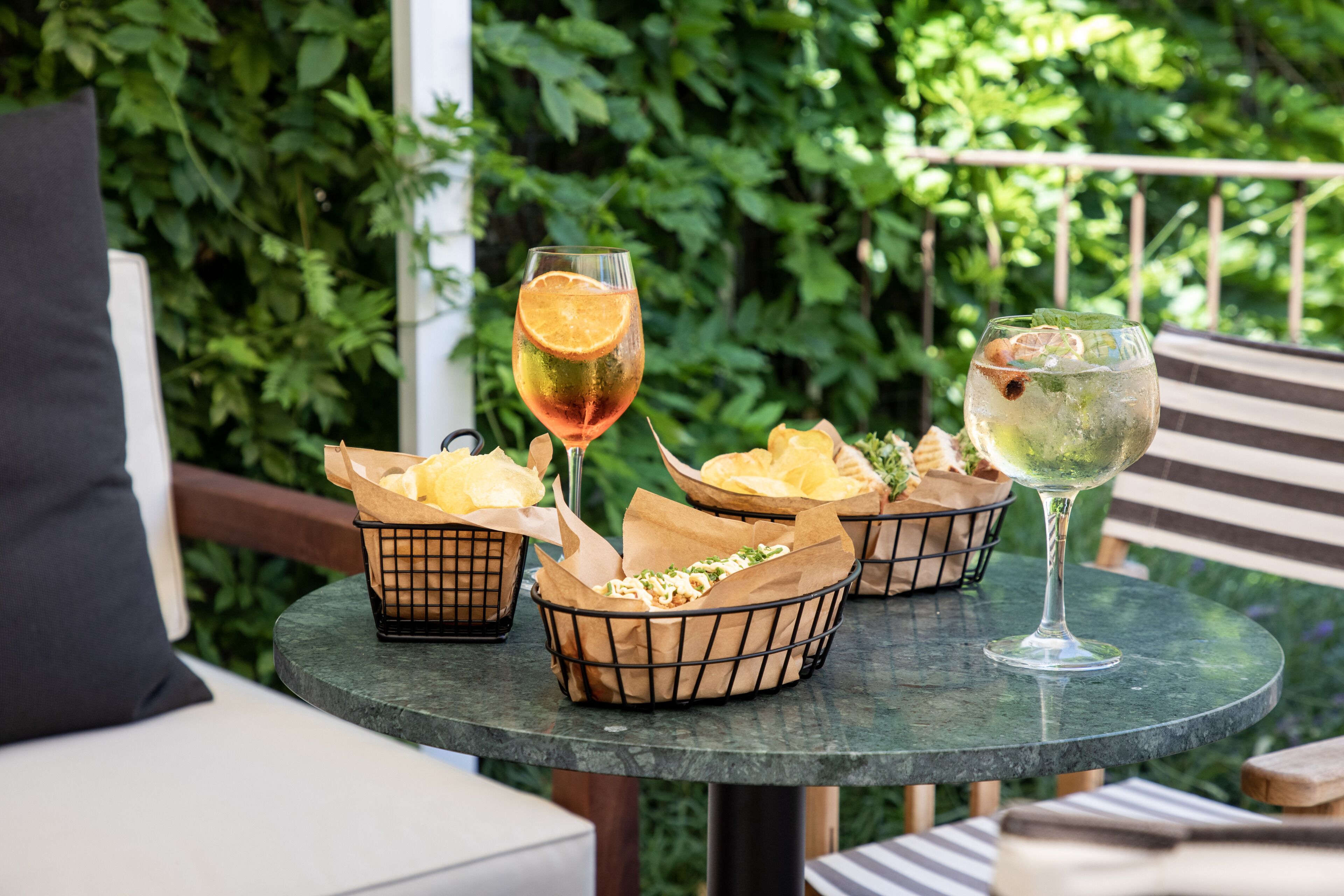 A table with baskets of food and two drinks, set outdoors with greenery in the background.