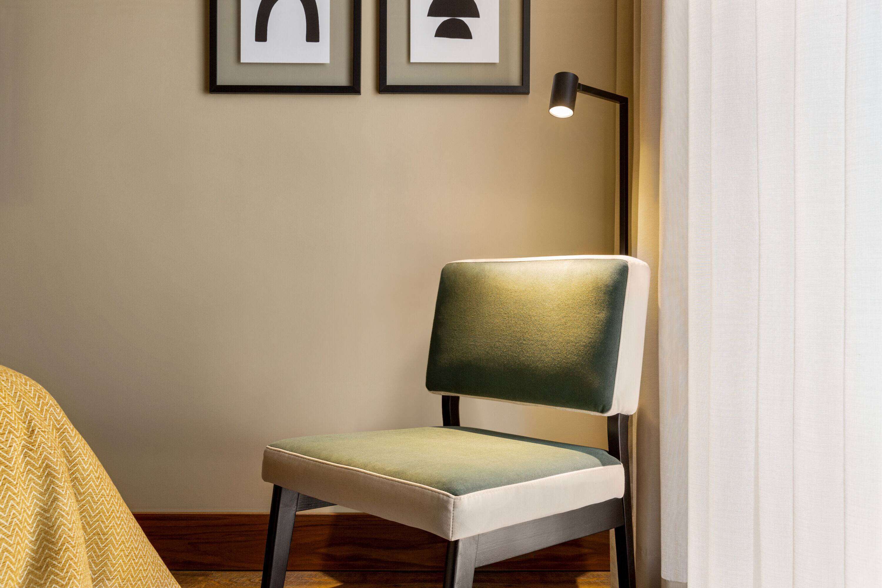 A chair with a cushioned seat and backrest, positioned next to a wall with framed artwork and a floor lamp.