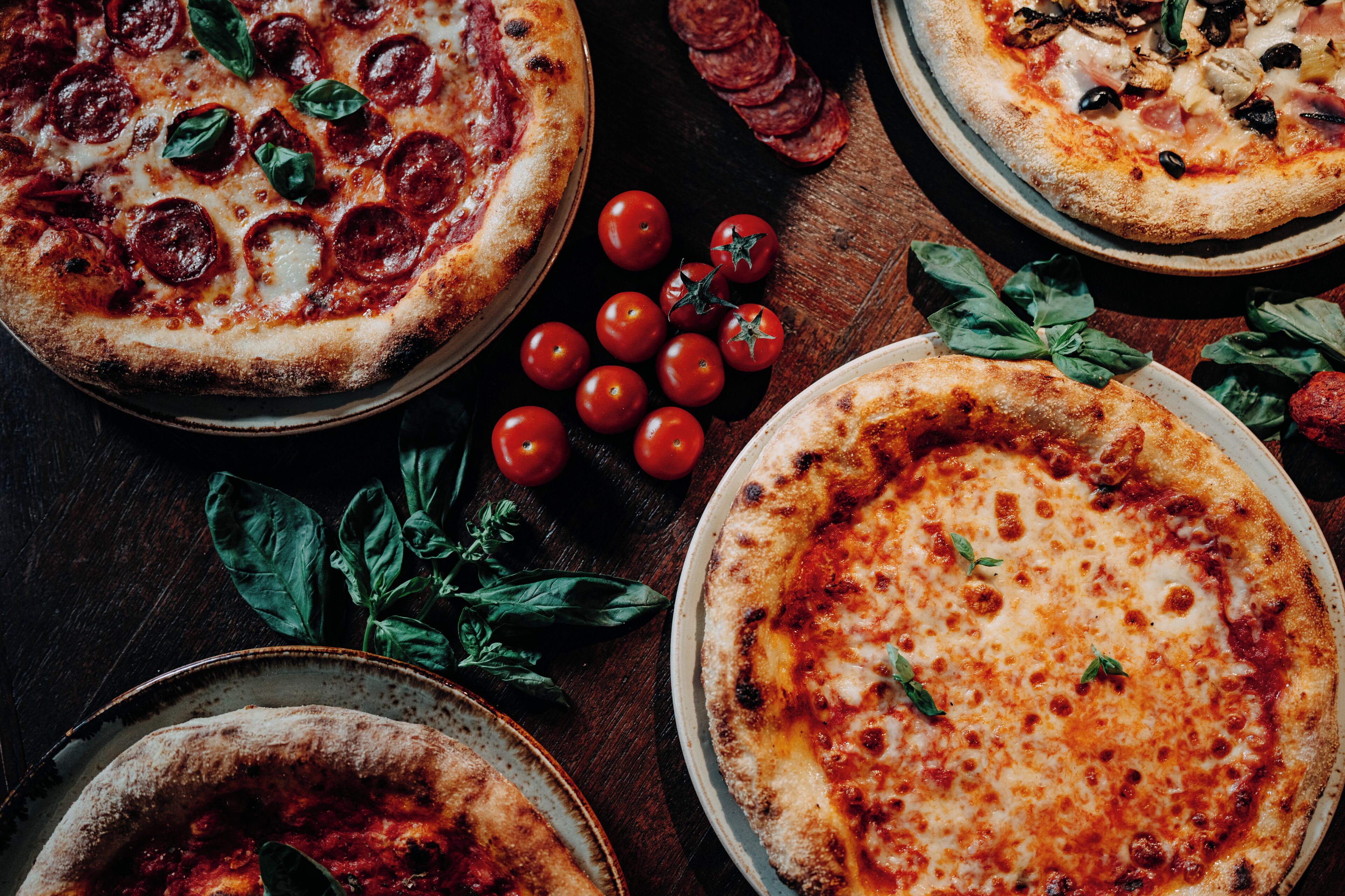Three pizzas with various toppings and fresh tomatoes on a wooden table.