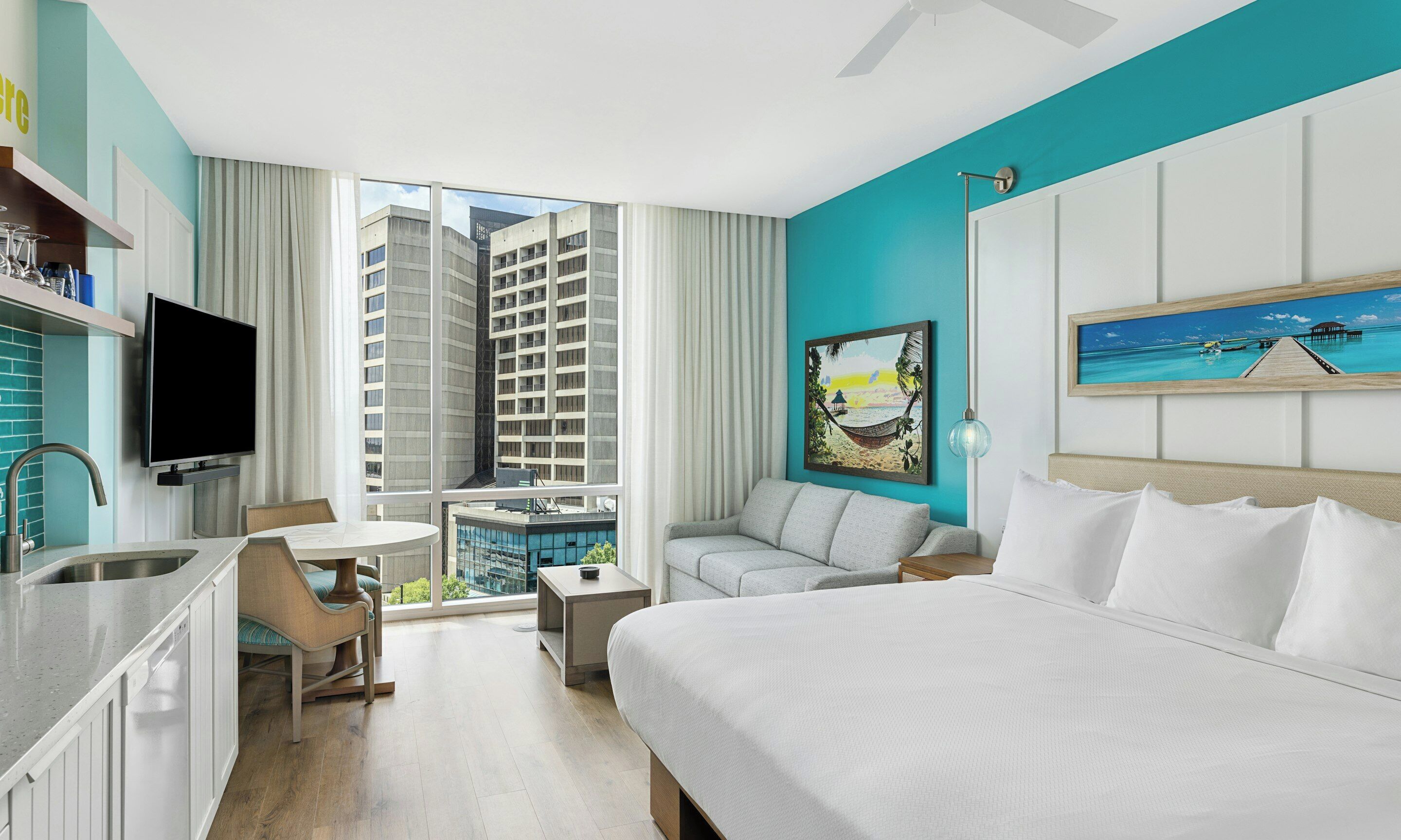 A hotel room with a bed, a TV, a desk, a chair, a window with a city view, and artwork on the wall.