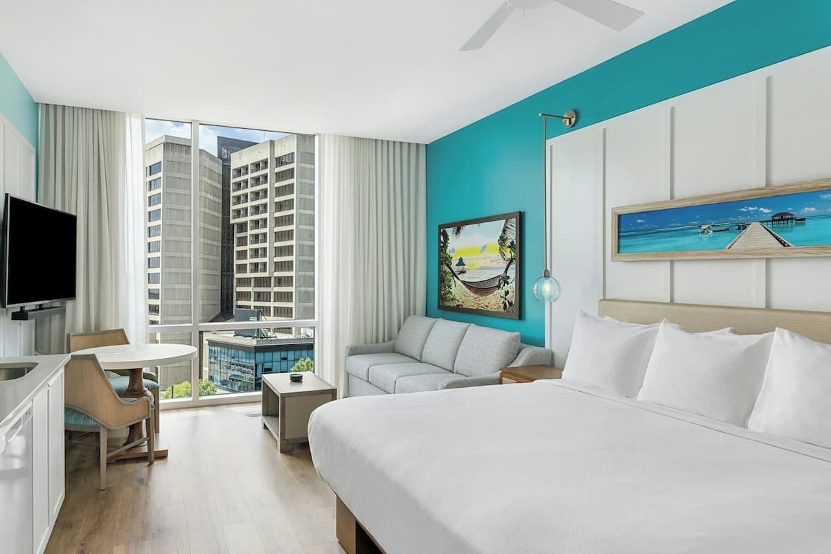 A hotel room with a bed, a TV, a desk, a chair, a window with a city view, and artwork on the wall.