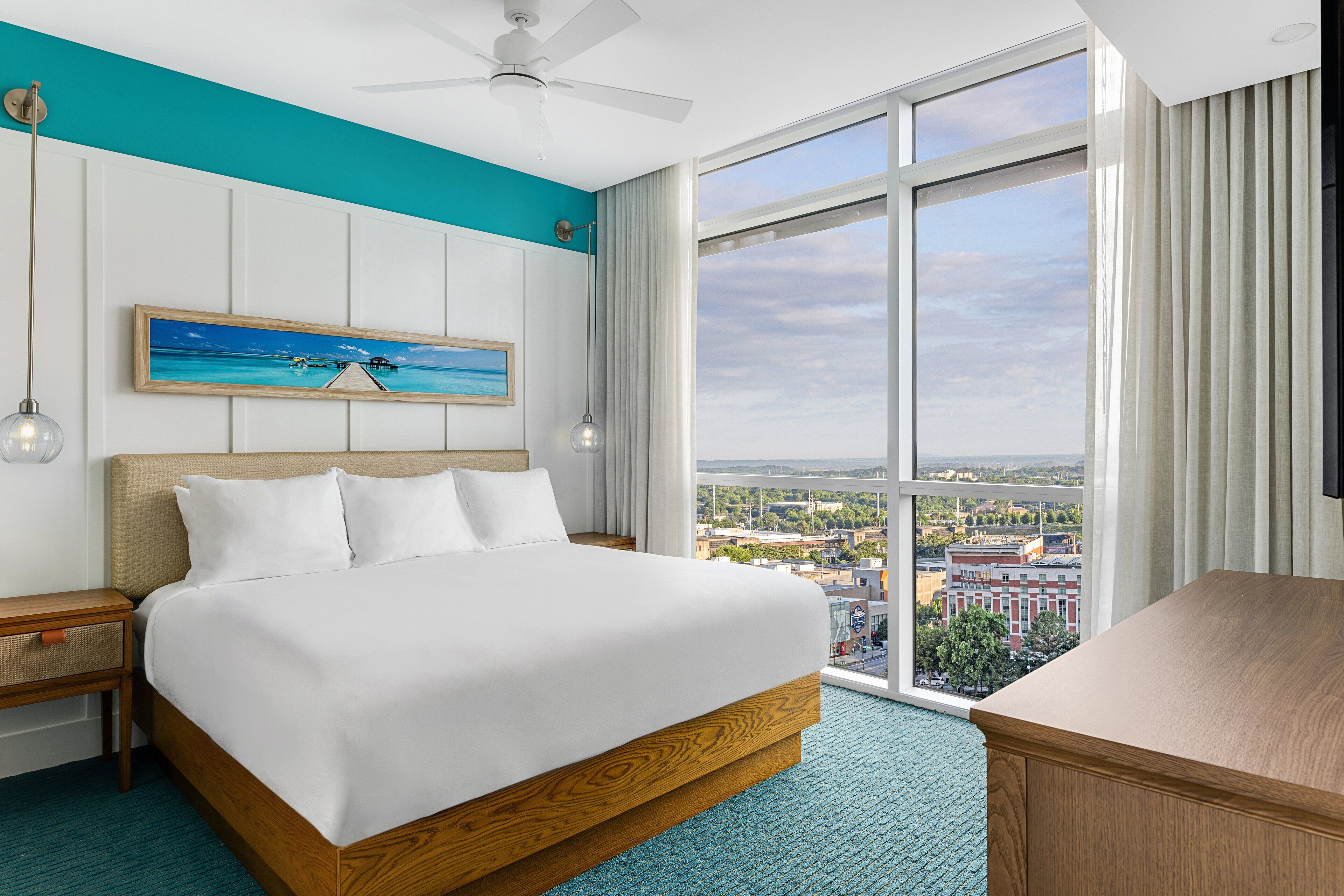 A hotel room with a large bed, a view of the city, a ceiling fan, and a desk.