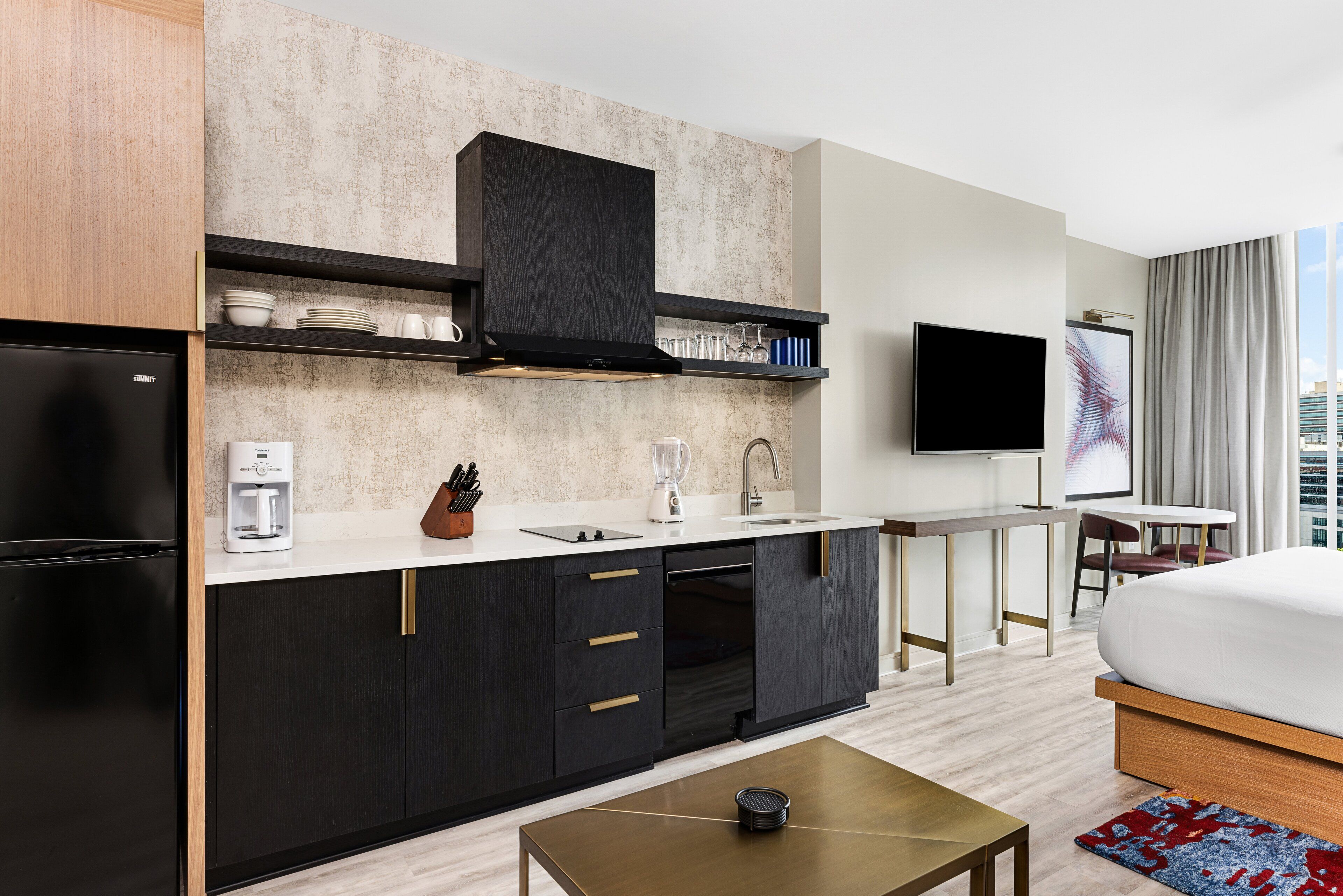A modern hotel room with a kitchen area, a bed, a TV, and a city view.