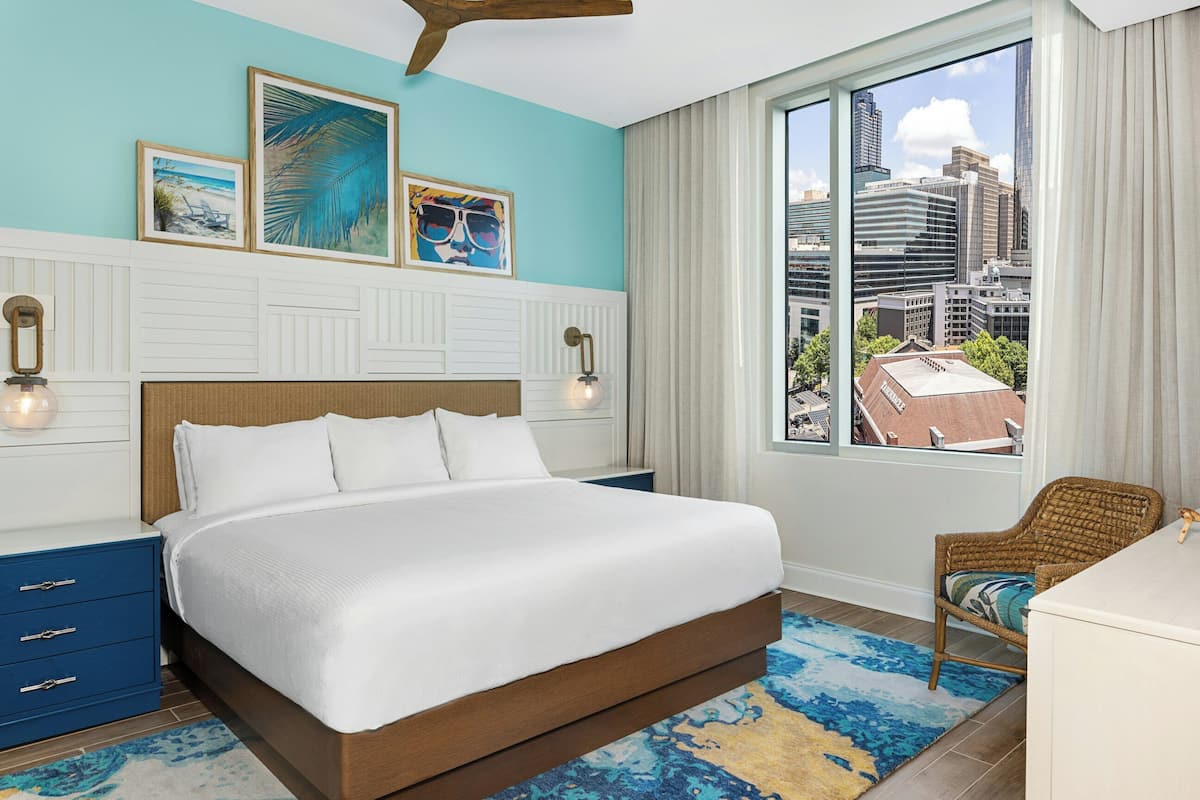 A hotel room with a bed, a chair, a desk, and a large window with a city view.