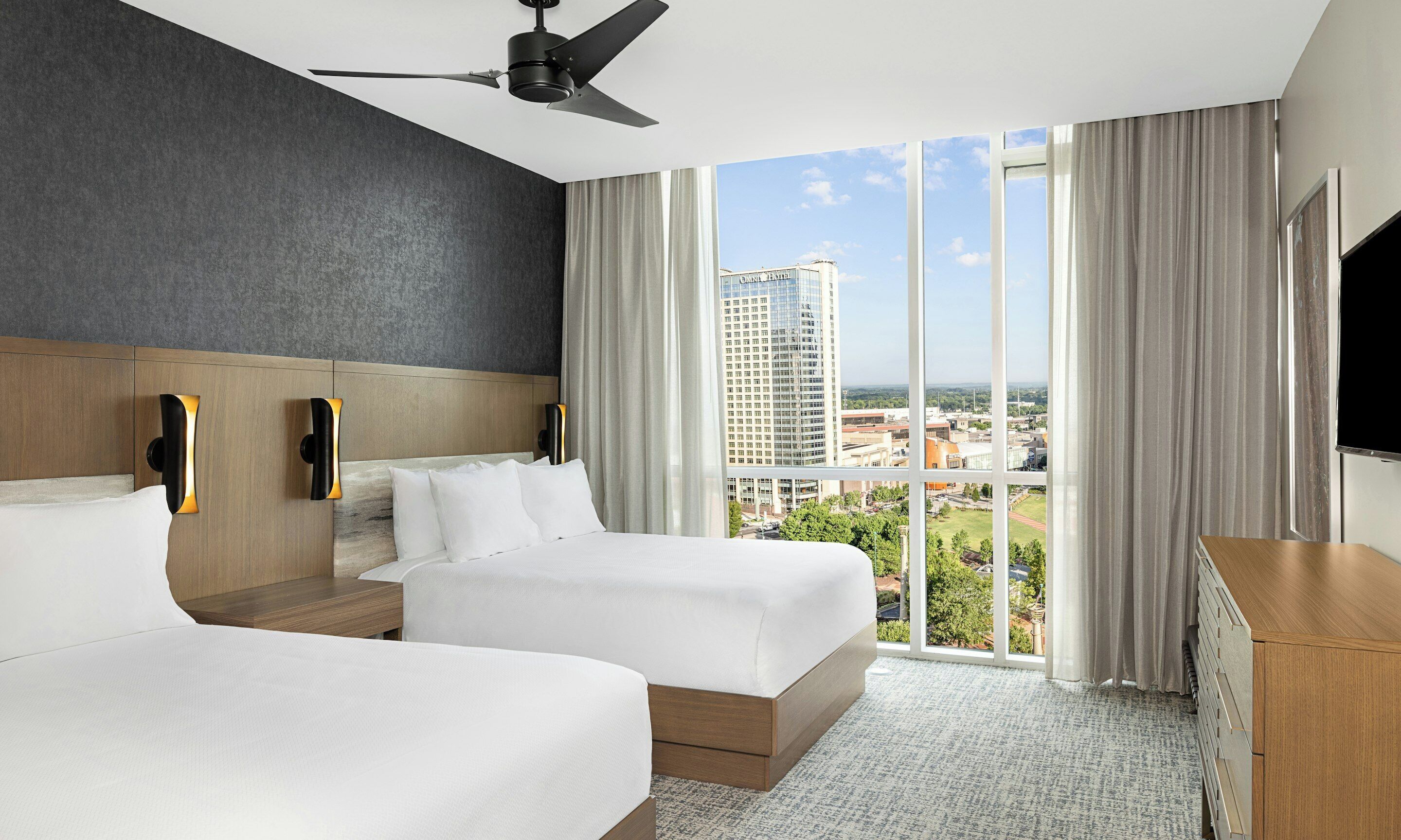 A hotel room with two beds, a ceiling fan, a TV, and a large window with a city view.
