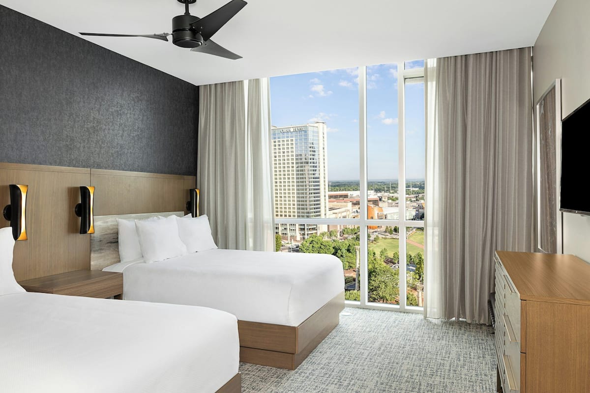 A hotel room with two beds, a ceiling fan, a TV, and a large window with a city view.