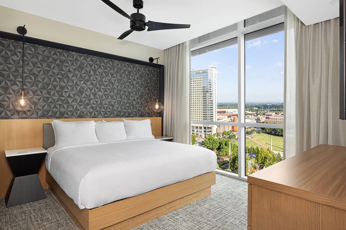A modern hotel room with a large bed, a desk, a ceiling fan, and a view of a cityscape through large windows.