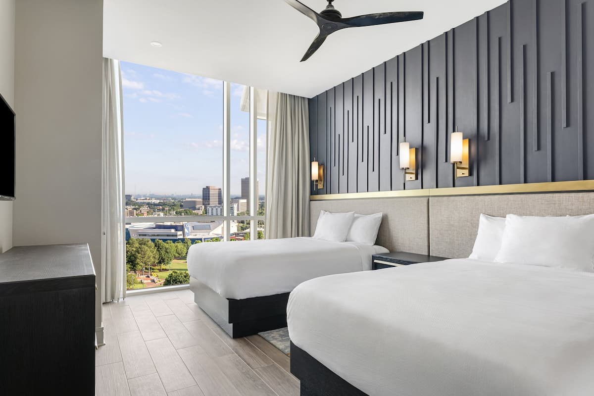 A hotel room with two beds, a ceiling fan, a large window with a city view, and a modern wall design.