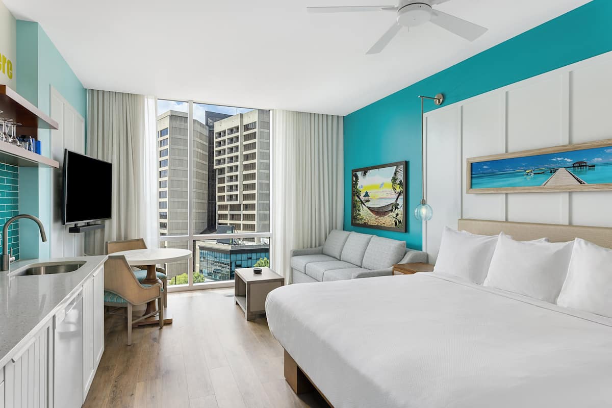 A modern hotel room with a large bed, a desk with a chair, a sofa, a TV, and a balcony with a view of tall buildings.