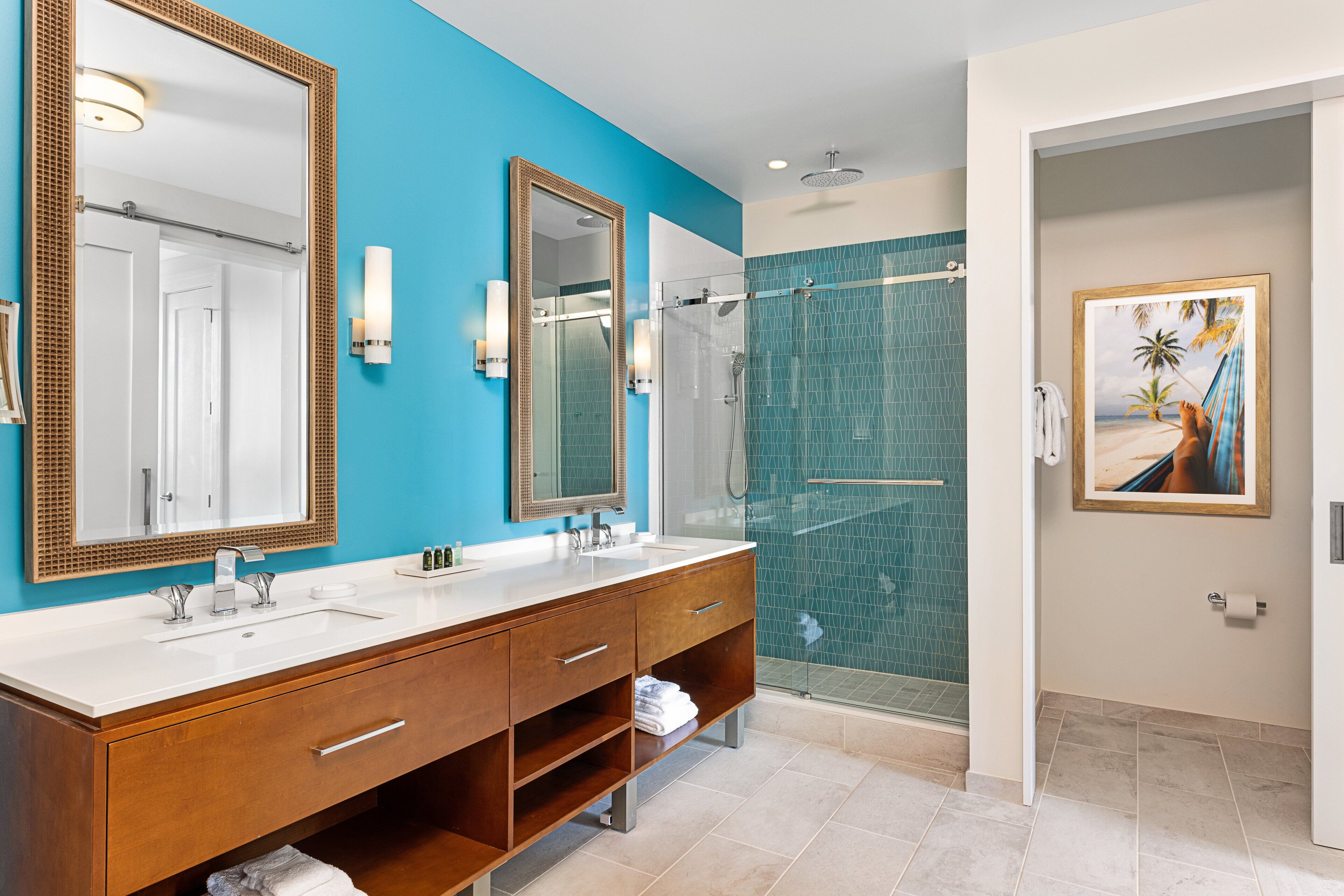 A bathroom with a blue accent wall, a large mirror, a wooden vanity, a glass shower enclosure, and a framed picture on the wall.