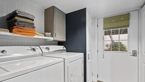 A laundry room with two front-loading washing machines, a stacked shelf with folded towels, and a door with a window view.