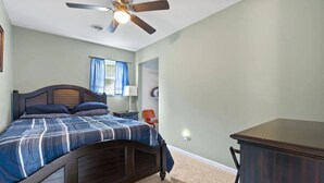 A bedroom with a bed, a desk, a chair, a ceiling fan, and a window with curtains.