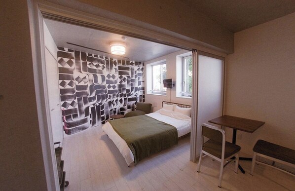 A bedroom with a bed, a bedside table, a chair, a wall with a geometric pattern, and a window with a view of greenery.