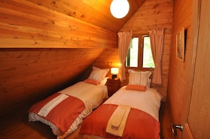 A cozy cabin room with two beds, wooden walls, a window with curtains, and a warm light fixture.