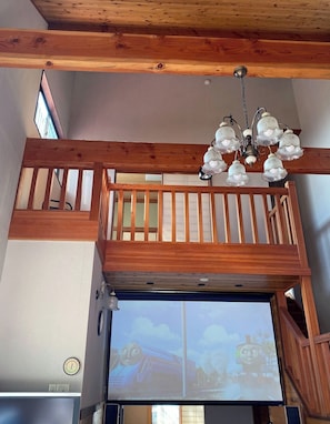 A multi-level wooden interior with a balcony, a chandelier, and a projection screen displaying a cartoon character.