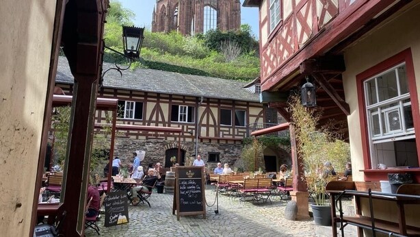Un café al aire libre con suelo de piedra, mesas y bancos de madera. Se ve una iglesia con una torre alta al fondo.