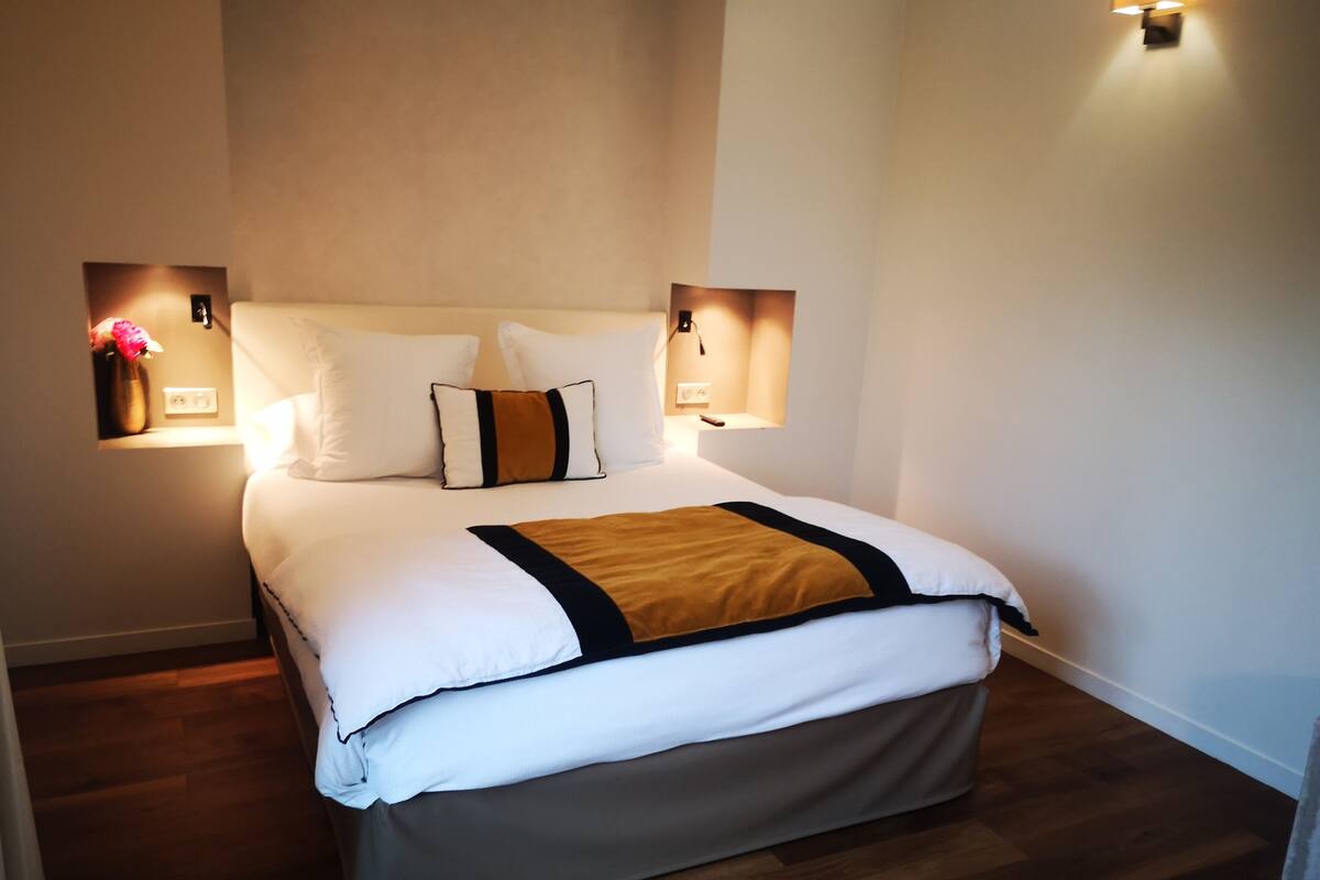 Una cama bien hecha con ropa blanca y un detalle en marrón, flanqueada por dos lámparas de pared.