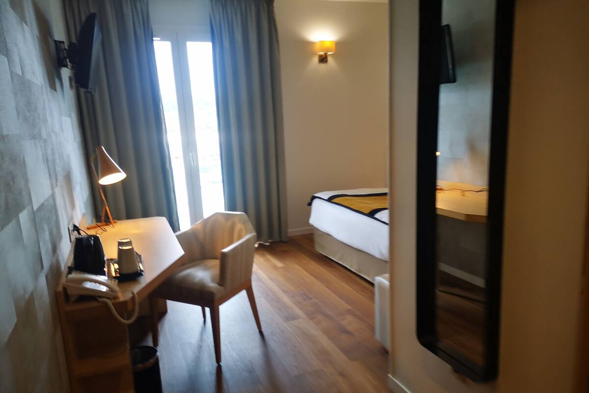 Una habitación de hotel con cama, escritorio, silla, lámpara y ventana con cortinas.