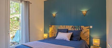 A bedroom with a large bed, two bedside lamps, a window with curtains, and a view of greenery outside.