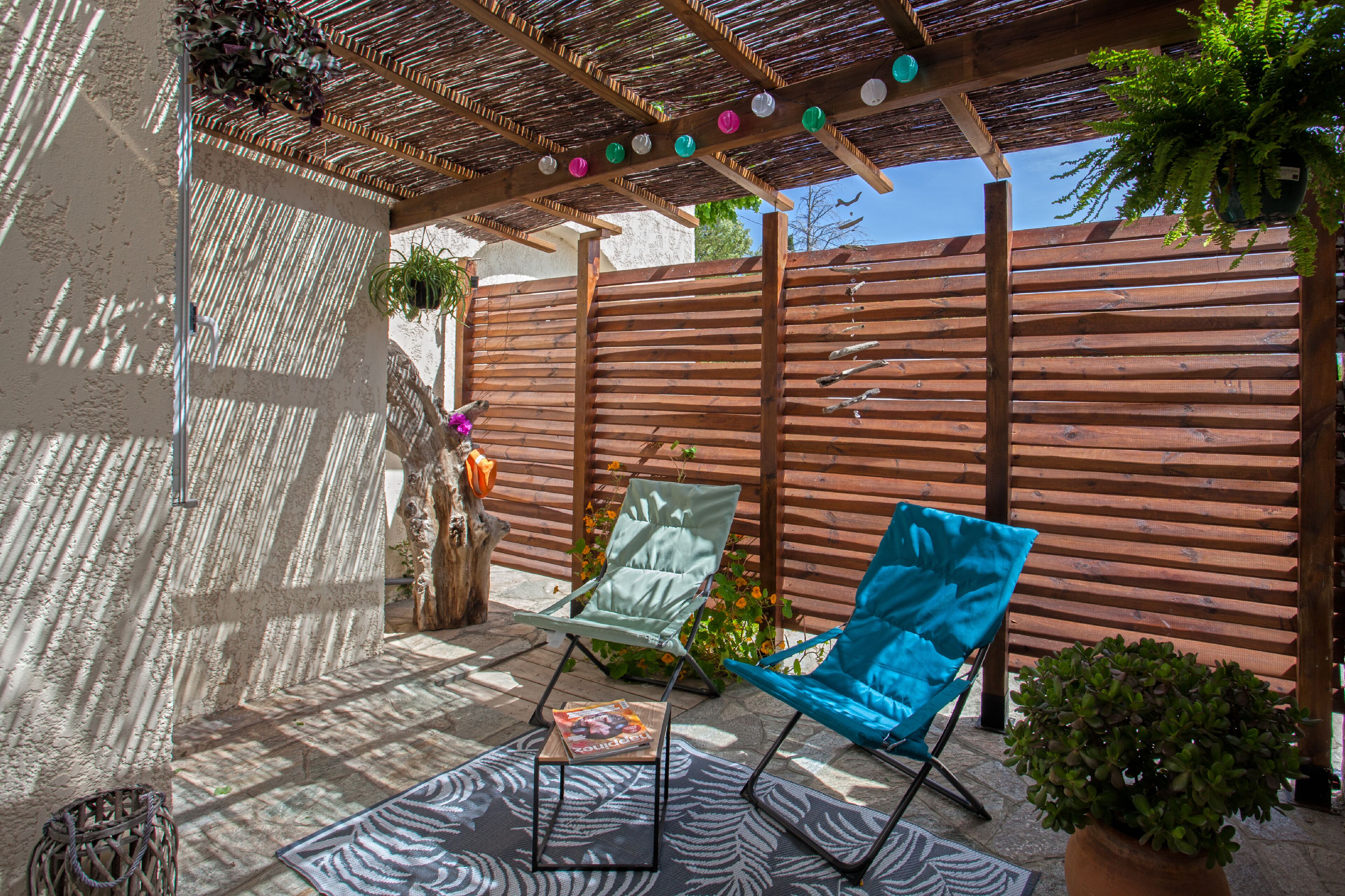 Een terras met een houten pergola, een blauwe stoel, een kleine tafel met een tijdschrift en een plant in een pot.