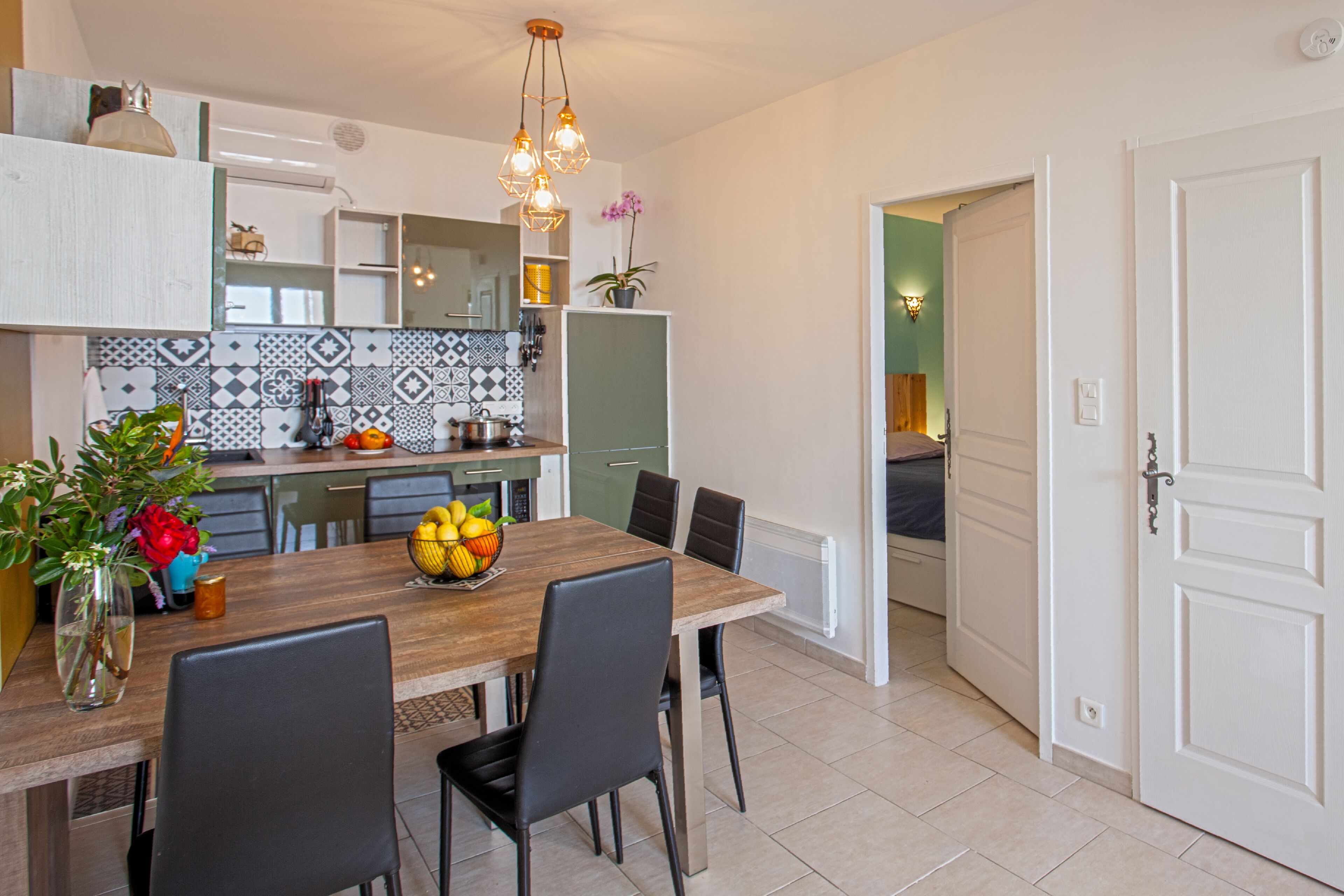 Een moderne keuken met een houten eettafel, zwarte stoelen en een fruitschaal. Op het aanrecht staat een potplant en boven de tafel hangt een lamp.